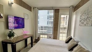 Ein Schlafzimmer mit einem großen Bett, einem an der Wand montierten Fernseher, einer Bank und einem Fenster mit Blick auf Gebäude.