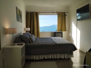 Un dormitorio con cama, mesita de noche, lámpara, ventana con cortinas y televisor en la pared.