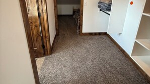 Ein schmaler Flur mit Teppichboden, einer Holztür und einem weißen Schrank mit Regalen und einer hängenden Dekoration.