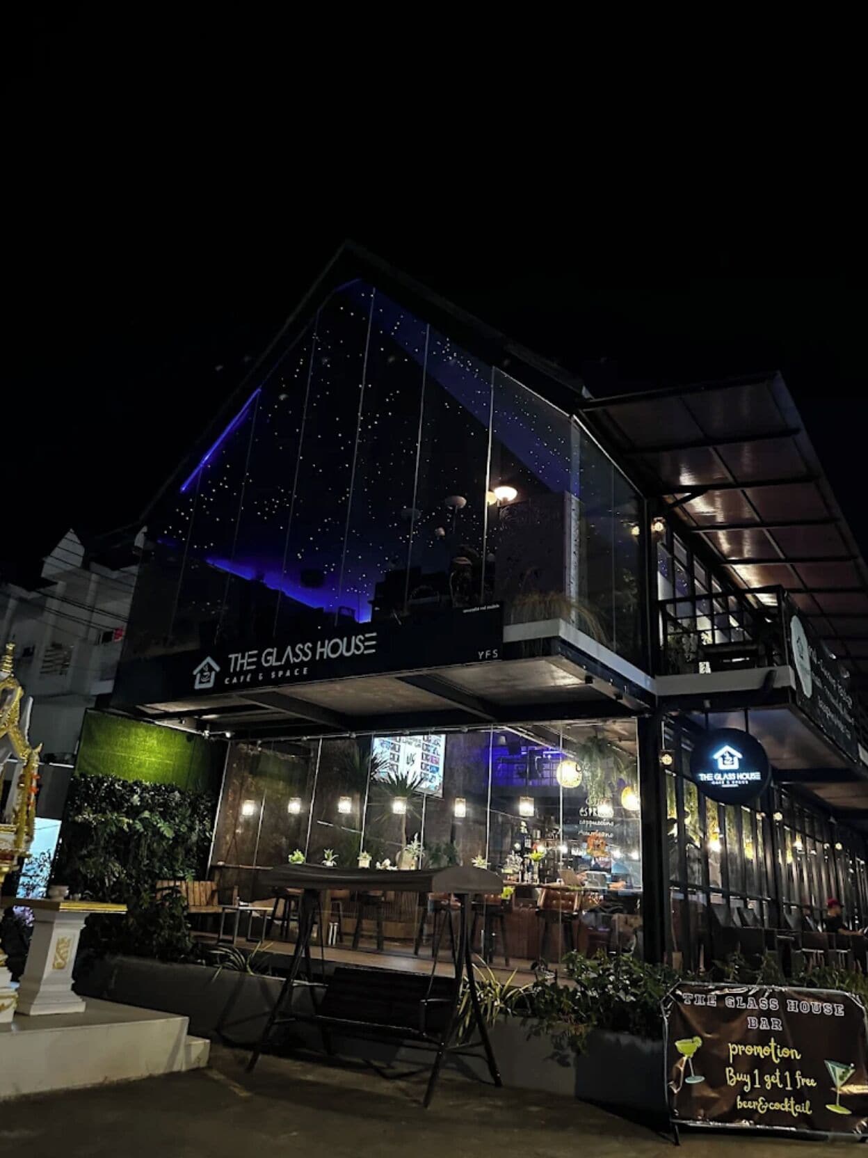 A modern building with a glass facade, labeled 'The Glass House,' featuring a bar area with outdoor seating and a sign offering a promotion.