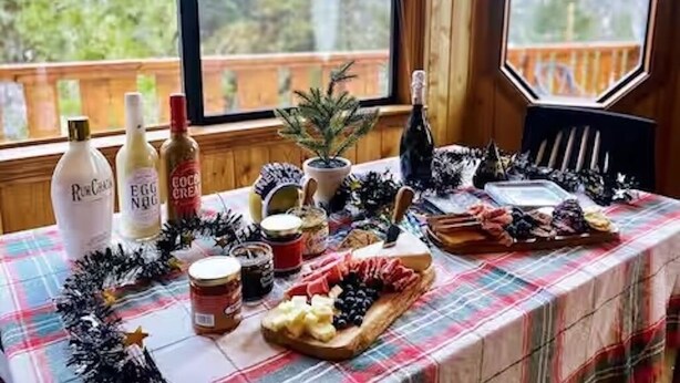 A table set with food and drinks, a Christmas tree, and a view of trees through a window. - L'Après Chalet · Bear Mtn Views · Sleeps 6 · Pet-Friendly (Big Bear Lake)