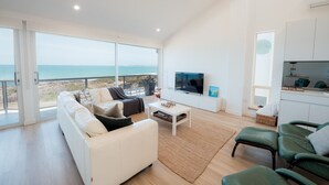 Uma sala de estar espaçosa com um sofá grande, uma mesa de centro e uma televisão. Há uma varanda com vista para o mar.
