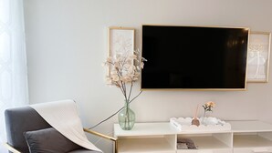 A modern living room with a grey sofa, a glass vase with flowers, a white console table, and a large flat-screen TV mounted on the wall. - 3 Bedroom Sunny Cape Coral Escape, 3 blocks from Lee Health, no cleaning fee! (Cape Coral)