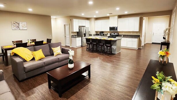A modern living room with a sofa, coffee table, and a dining area with chairs. A kitchen with white cabinets and appliances is visible in the background. - Fabulous 4-bedroom house with AC in Riverside (Riverside)