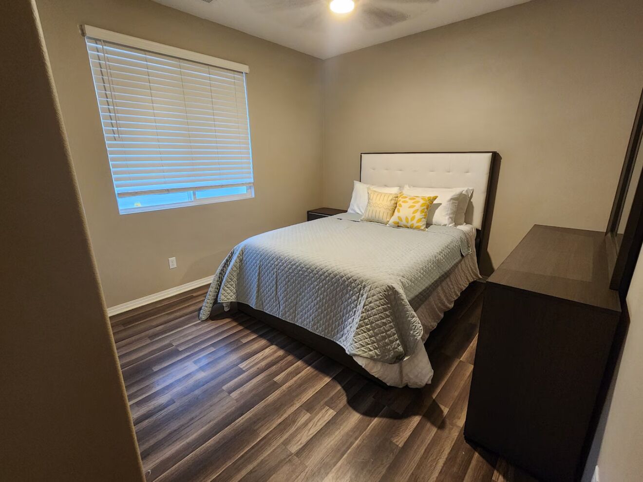 Une chambre à coucher comprenant un lit, une table de chevet, une fenêtre avec des stores et une commode de couleur foncée.