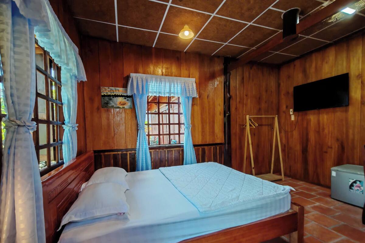 A wooden room with a bed, a TV mounted on the wall, and a window with curtains.
