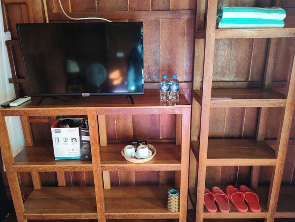 A wooden TV stand with a flat-screen TV, a remote control, two water bottles, a box of instant coffee, a tray with cups, and a pair of red slippers. - Rooster Mekong Resort (Cho Lach)