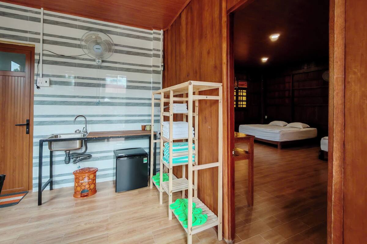 A compact room with a kitchenette, a bed, and a wooden shelf unit.