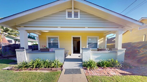 A single-story house with a yellow facade, a front porch with two planters, and a clear pathway leading to the entrance.