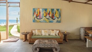 A room with a wicker sofa, a patterned ottoman, a painting of sailboats, and a view of the ocean. - Naluna Glamping Lembongan (Lembongan Island)