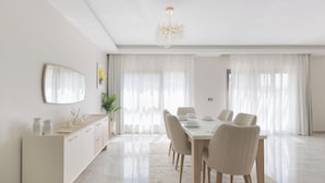 Una sala da pranzo moderna con un tavolo bianco imbandito per sei persone, circondato da sedie beige. Un grande specchio è appeso alla parete, e una lampadario è visibile sopra il tavolo.