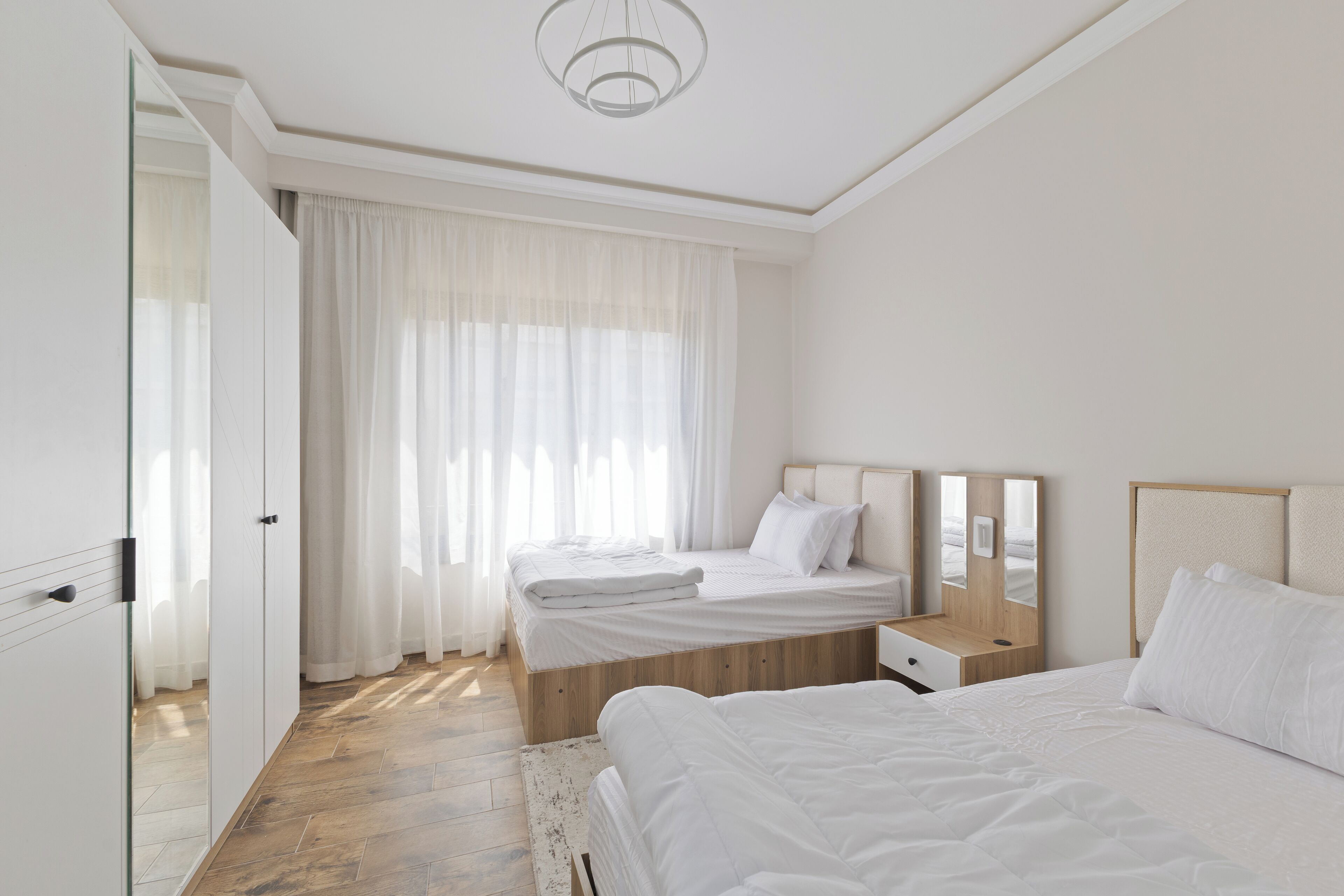 A modern hotel room with two beds, a large window with sheer curtains, a wooden floor, and a minimalist design.