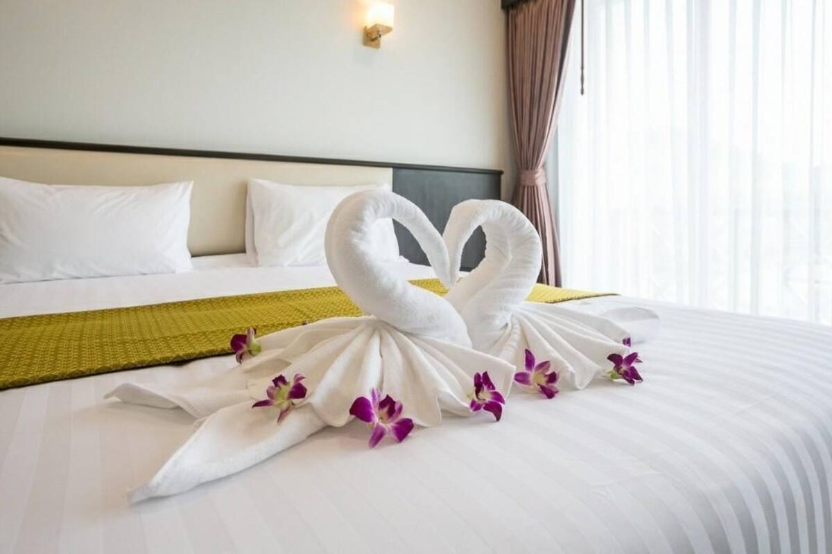 Un lit bien fait, agrémenté de deux serviettes en forme de cygne et d’orchidées violettes, d’un plaid jaune et de coussins blancs.