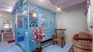 Una habitación con un separador azul, una silla de madera, una bañera de madera y un florero con flores.