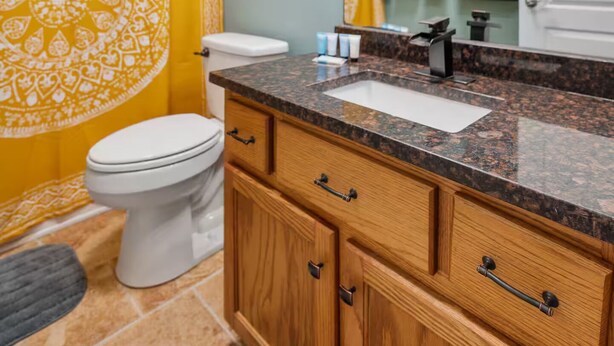 A bathroom with a yellow patterned shower curtain, a white toilet, a granite countertop sink, wooden cabinets, and a mirror above the sink. - Spacious, Pristine Home w/ Firepit, Bourbon Lounge (Bardstown)