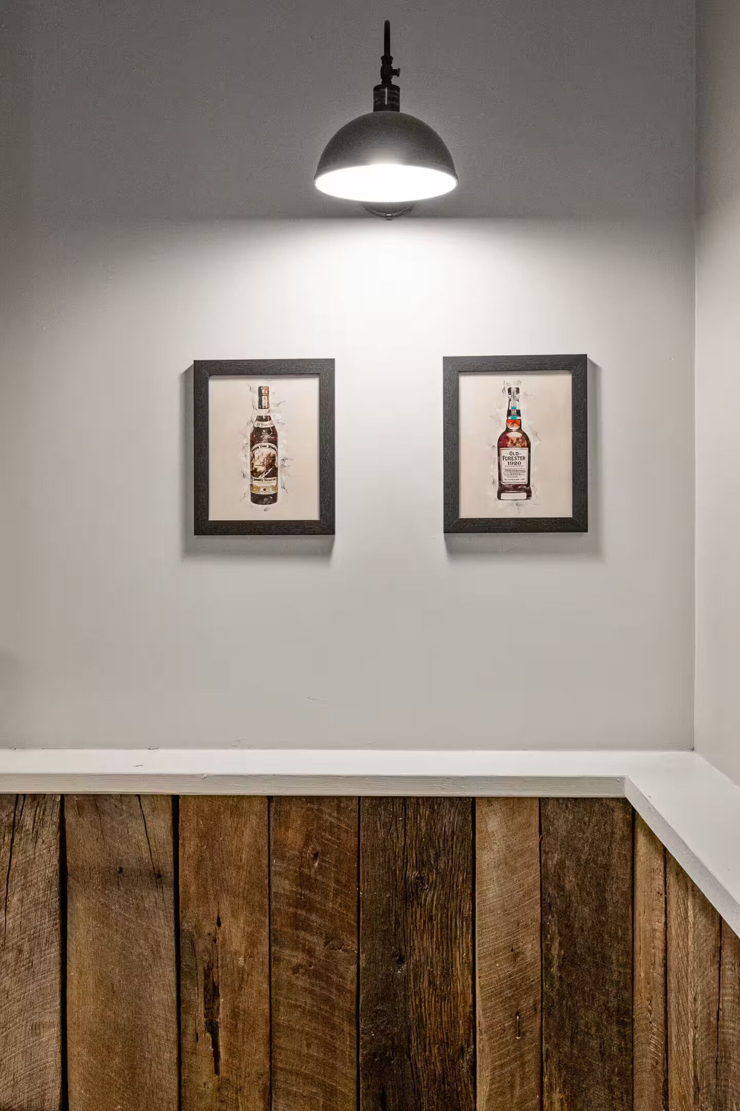 Two framed pictures of a bottle on a white wall, under a single light fixture.