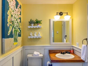 A bathroom with a wooden vanity, a white sink, a large mirror, and a painting of flowers on the wall. - Historic Maine Cottage with Wrap-Around Porch & Ocean Views (Boothbay)