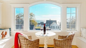 Een kamer met een groot rondboogvenster, rieten stoelen, een tafel met fles en glas, en uitzicht op zee en gebouwen.