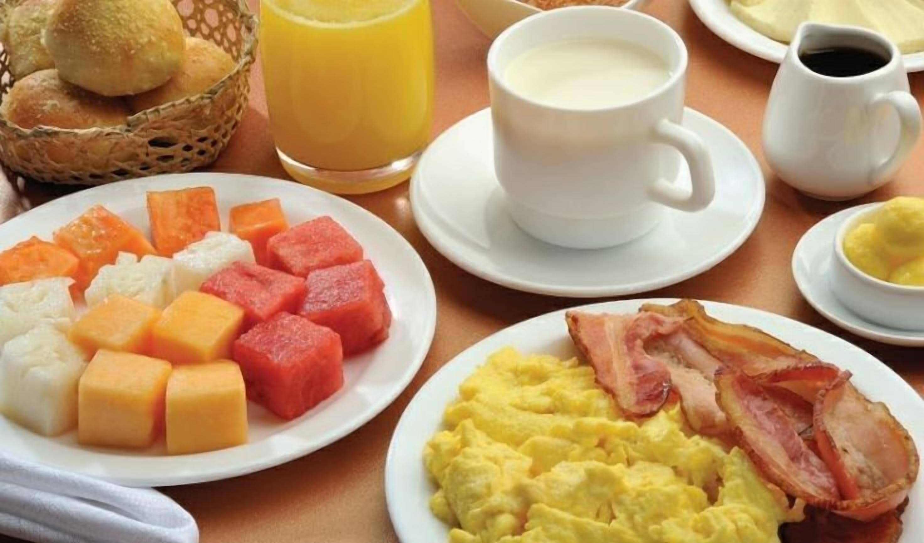 A breakfast setting with plates of eggs, bacon, and fruit, a cup of milk, and a glass of orange juice.