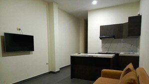 A compact living space with a flat-screen TV mounted on the wall, a kitchenette with dark cabinets, and a brown sofa.
