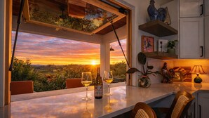 Uma área de refeições com vista para um pôr do sol sobre um vinhedo, uma garrafa de vinho e dois copos na mesa.