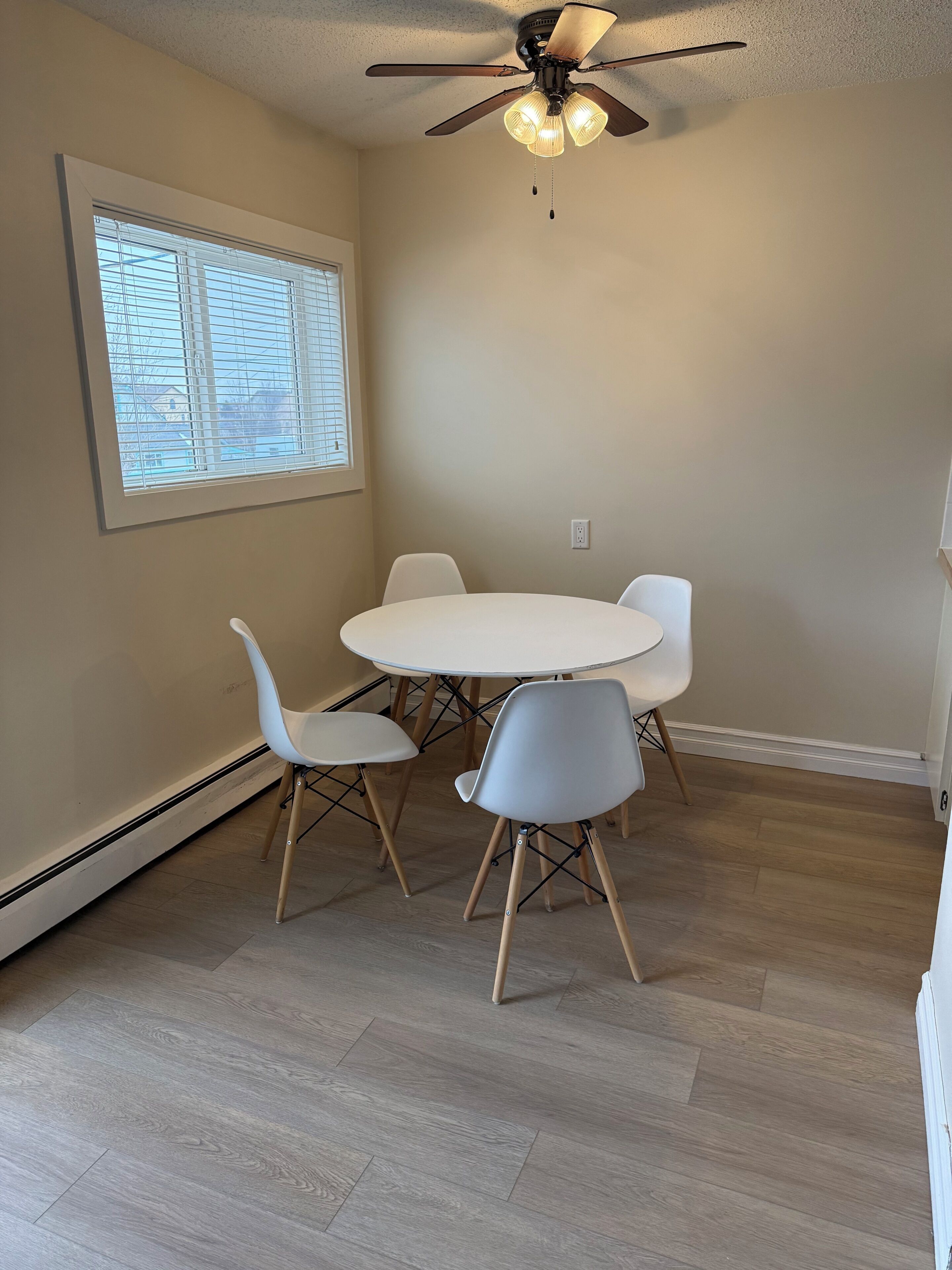 Une pièce avec un ventilateur de plafond, une table ronde entourée de quatre chaises, une fenêtre avec des stores et un interrupteur sur le mur.