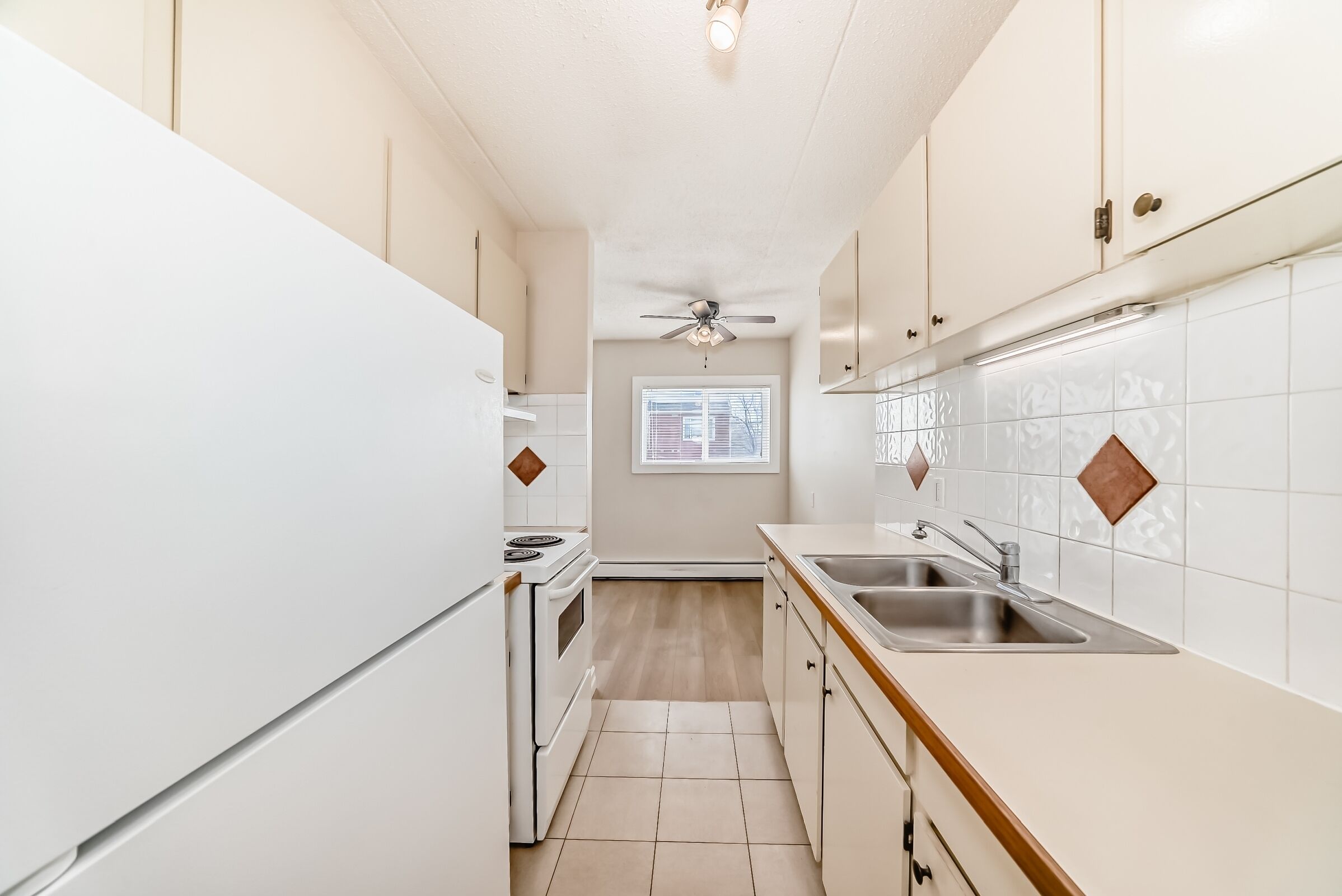 A narrow kitchen with white cabinets, a stove, a sink, and a window with blinds.