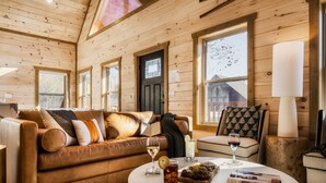 Ein gemütliches Blockhaus-Interieur mit einem braunen Ledersofa, einem runden Couchtisch mit Getränken und Snacks sowie großen Fenstern mit Blick ins Freie.