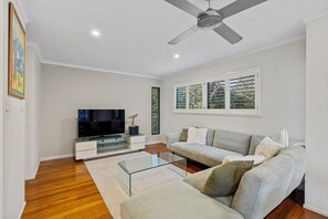 A modern living room with a flat-screen TV, a glass coffee table, a grey sofa, and a ceiling fan.