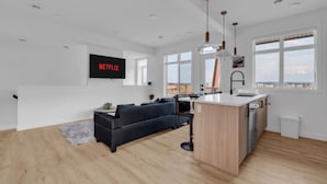 A modern living room with a black sofa, a wooden dining area, and a large window with a view. - Modern 3BR Townhome in Seton with Garage + Balcony (Calgary)