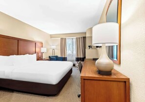 A hotel room with a large bed, a wooden nightstand, a desk, and a lamp.