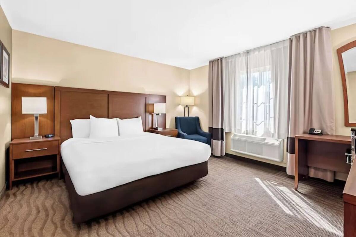 A hotel room with a large bed, a desk, a chair, a lamp, and a window with curtains.
