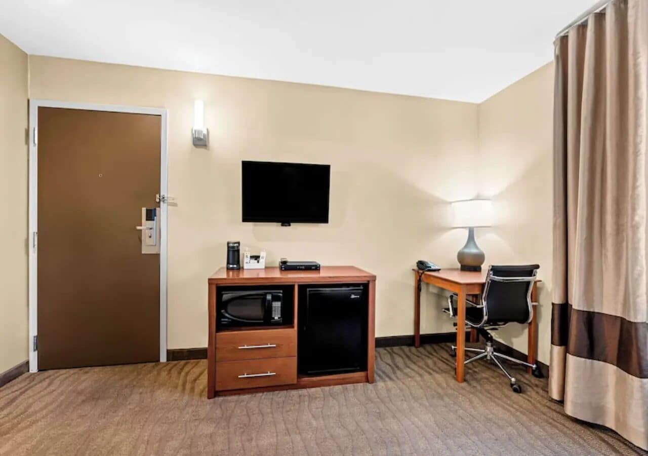 A hotel room with a wooden desk, a chair, a television mounted on the wall, a minibar, and a door.
