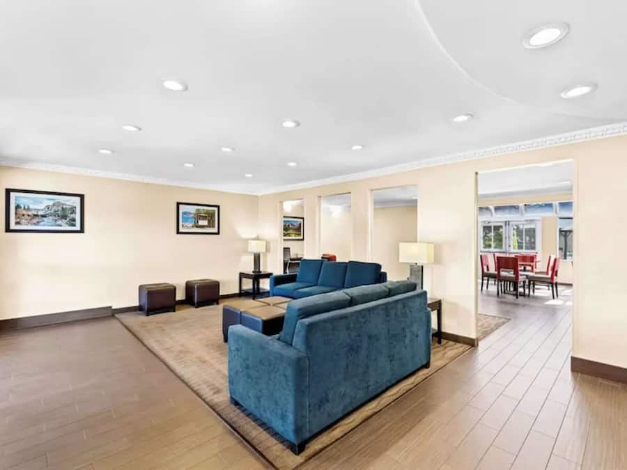 A spacious living room with a large blue sectional sofa, a small coffee table, and a rug. There are two side tables with lamps. The room has wooden flooring and a high ceiling with recessed lighting.