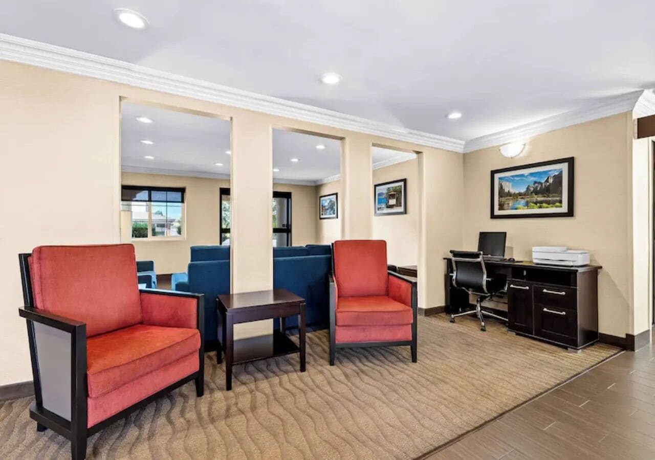 A hotel lobby with red armchairs, a small table, a blue sofa, a desk with a computer, and framed pictures on the wall.