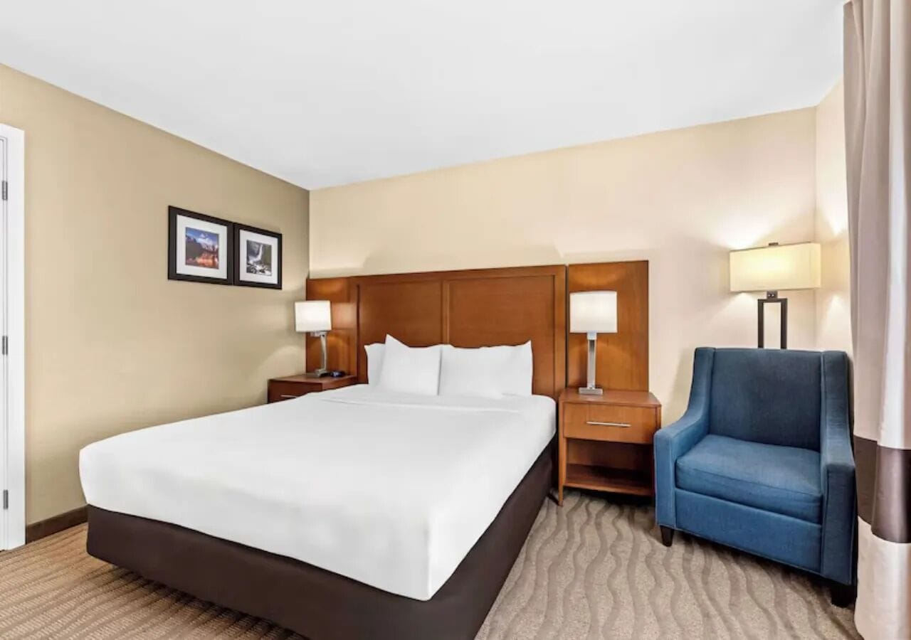 A hotel room with a large bed, two bedside tables with lamps, a blue armchair, and a wooden headboard.