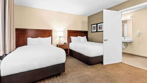 Una habitación de hotel con dos camas, una mesita de noche, una lámpara, un baño con lavabo y espejo, y una puerta que da a otra habitación.