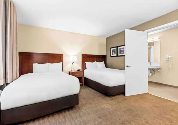 Una habitación de hotel con dos camas, una mesita de noche, una lámpara, un baño con lavabo y espejo, y una puerta que da a otra habitación.