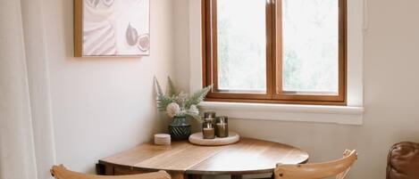 A dining area with a wooden table and two wicker chairs, a window with blinds, and a framed artwork on the wall.