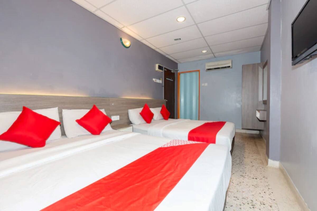 Hotellihuone, jossa on kolme sänkyä. Jokaisessa sängyssä on valkoiset vuodevaatteet ja punaiset koriste-elementit. Huoneen katossa on upotetut valaisimet ja seinälle on asennettu televisio.