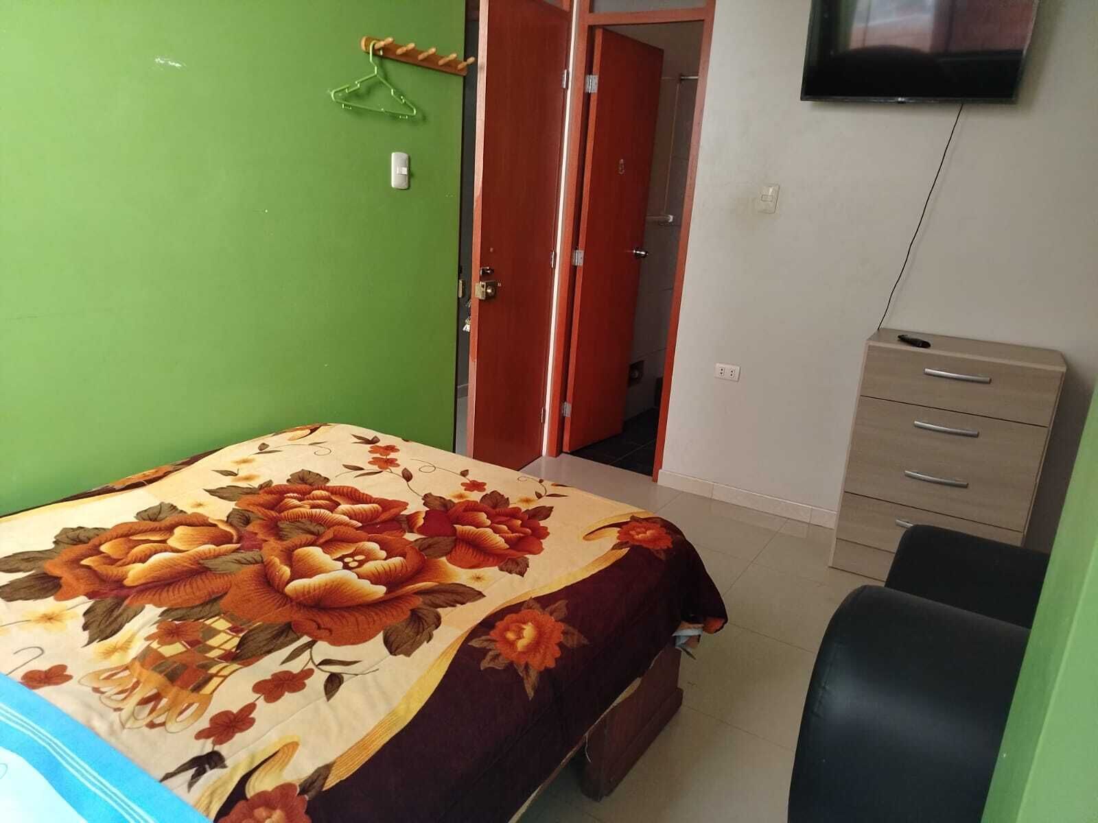 A bedroom with a floral bedspread, a wooden wardrobe, a chair, and a television on the wall.