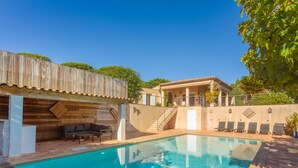 Una piscina con muelle de madera, zona de estar cubierta y una casa con techo de tejas al fondo.