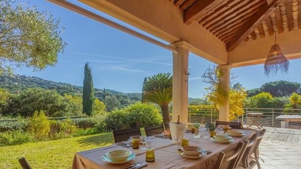 Un área de comedor al aire libre con una mesa dispuesta para cuatro personas, rodeada de sillas. La mesa está cubierta con un mantel color beige, y hay platos, cubiertos y vasos sobre ella. También se ve una botella de aceite de oliva. El entorno es exterior, con vistas a zonas verdes y colinas al fondo.