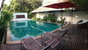 A swimming pool with wooden deck chairs and an umbrella, surrounded by greenery and a white wall. - Room-nakara Samui-superior Beach Front (Koh Samui)