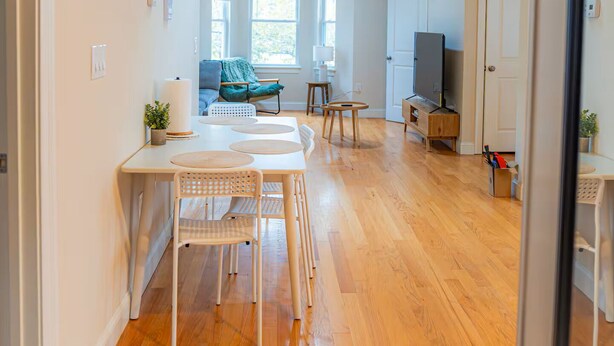 Een ruime woonkamer met een houten vloer, een eettafel met stoelen, een bank, een televisie en een deur naar een andere kamer.
