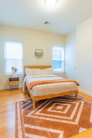 A bedroom with a wooden bed, a patterned rug, and two windows with blinds.