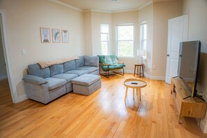 A living room with a sectional sofa, a green armchair, a small round table, and a television on a wooden stand.