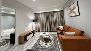 Ett modernt hotellrum med en säng, en soffa, ett soffbord, en bänk, en golvlampa och ett stort fönster med gardiner.