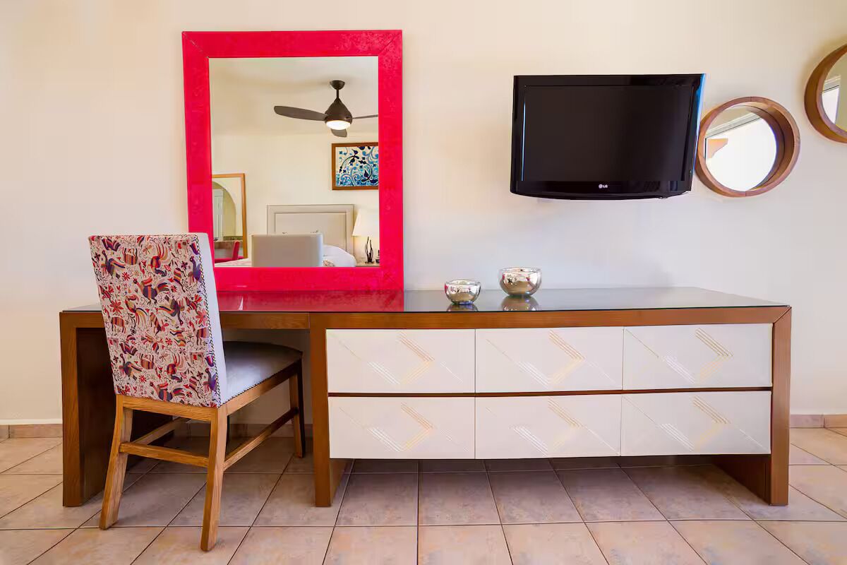 A hotel room with a wooden desk, a red-framed mirror, a flat-screen TV, and a chair with a patterned cushion.