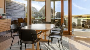 Een moderne eetruimte met een grote houten tafel en zwarte stoelen, grote ramen met uitzicht op een huis en landschap, en een hangende lamp.